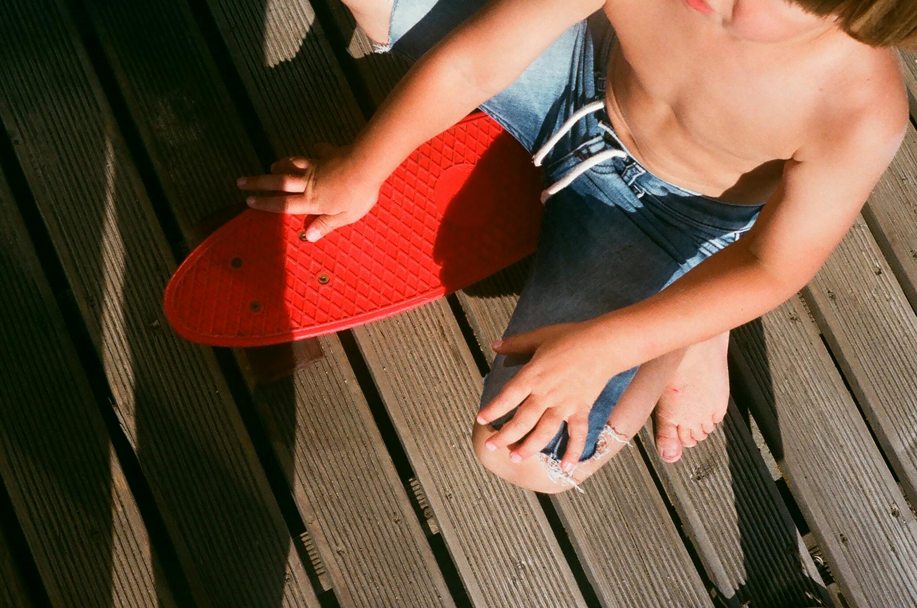 Dziecko w jeansowych spodenkach siedzi z czerwonym penny boardem na tarasie